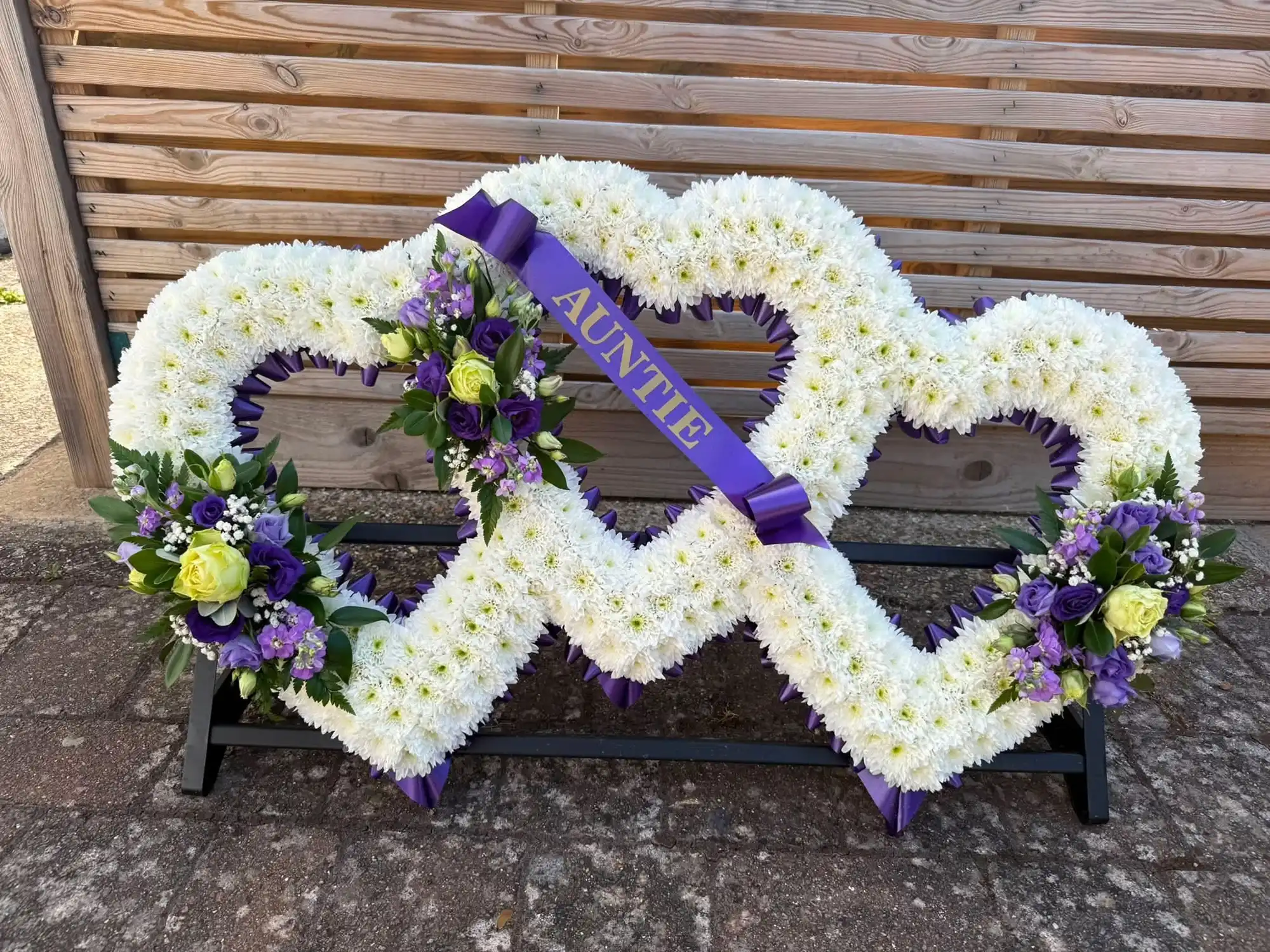 Double heart white floral funeral tribute with purple and yellow flower accents and a purple ribbon labeled Auntie.