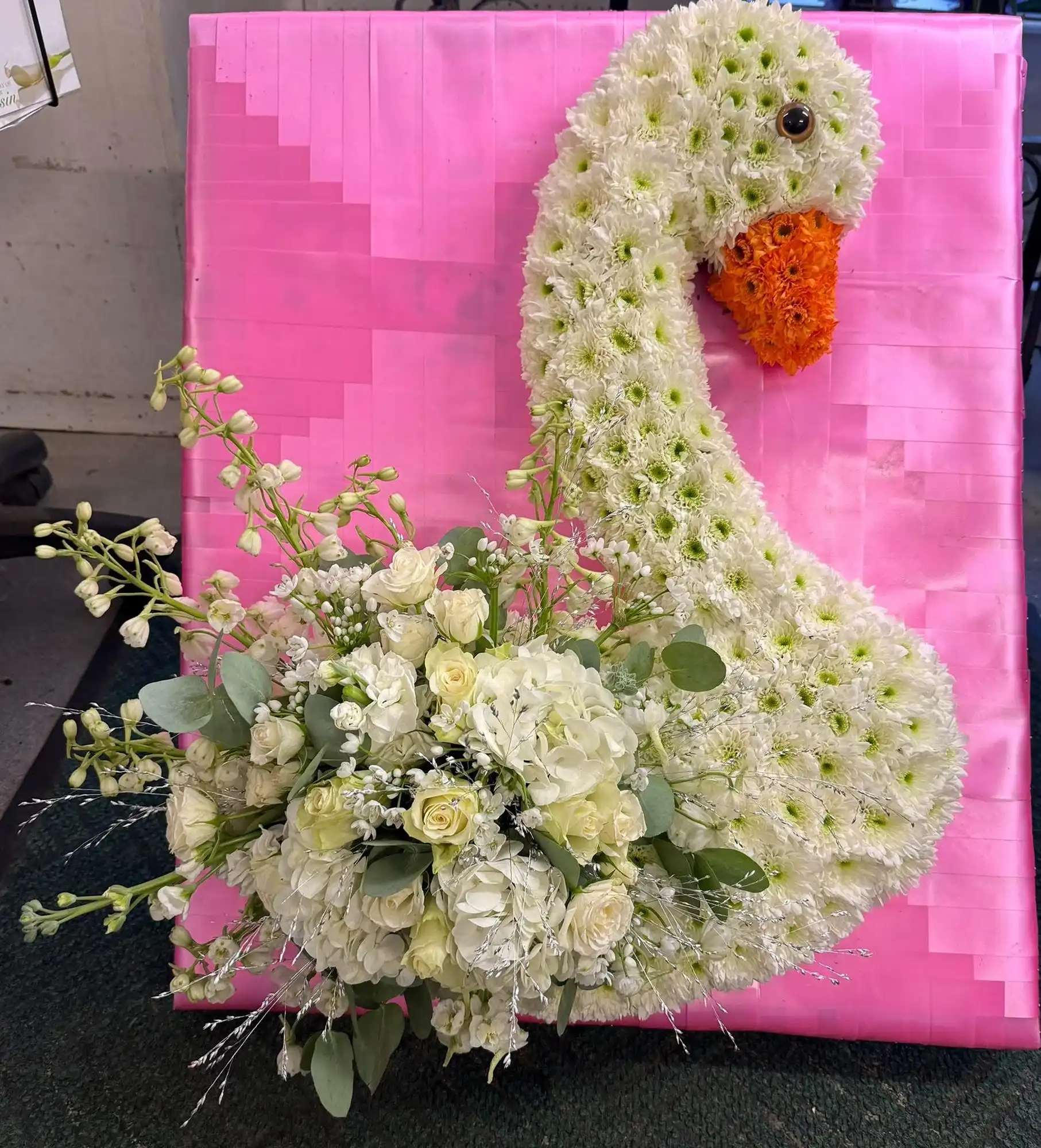 White flower arrangement shaped like a swan with an orange beak on a pink background.