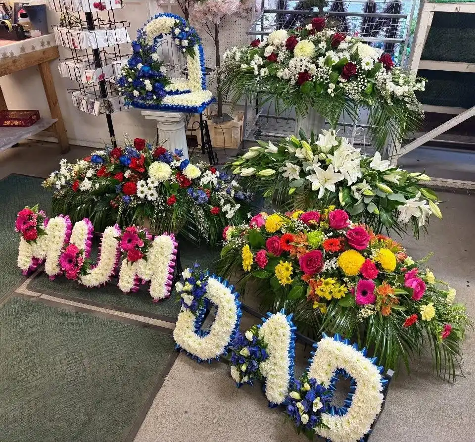 Multiple colorful funeral flower arrangements including letters spelling MUM and DAD in a shop.