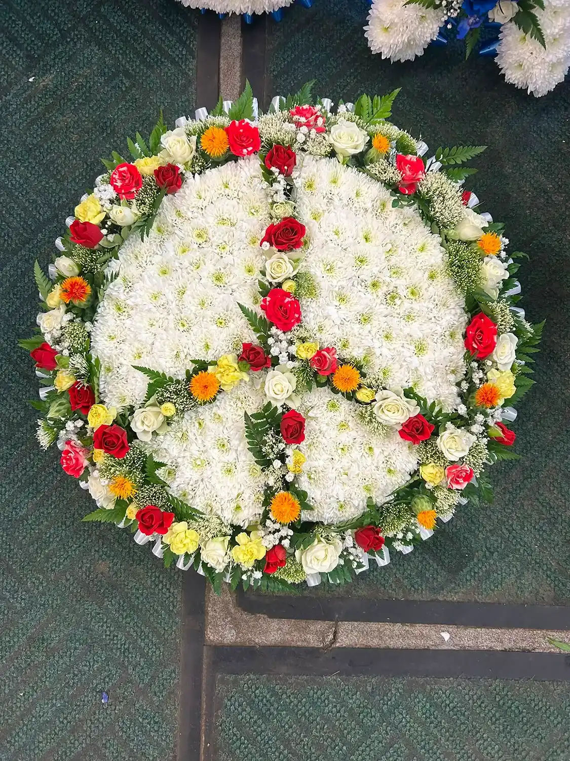 Round floral wreath with white flowers and a peace symbol made of red, yellow, and orange flowers.